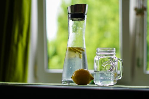 Lemon-infused water in a glass carafe and mason jar on a windowsill, evoking freshness.
