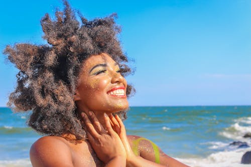 A joyful woman with afro hair smiling brightly on a sunny beach day.