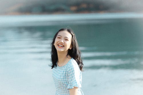 A cheerful young woman in a plaid dress laughing by a serene lake.