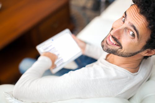 Happy man sitting on couch using a digital tablet at home, looking at camera.