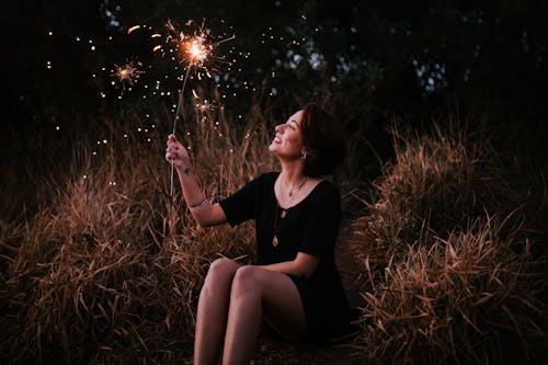 A smiling woman enjoying sparklers outdoors in a grassy field at night, expressing pure joy.