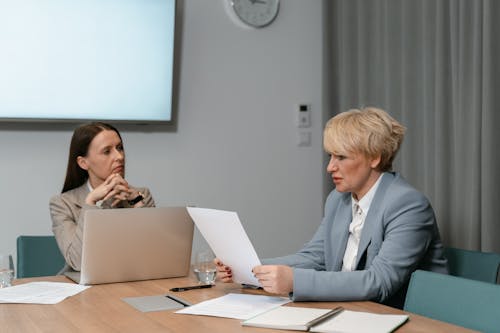 Two businesswomen in a serious meeting, discussing documents with a laptop in an office setting.